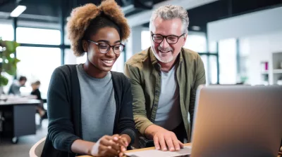 Experienced senior mentor guiding a smiling young professional with a laptop at a contemporary office space.